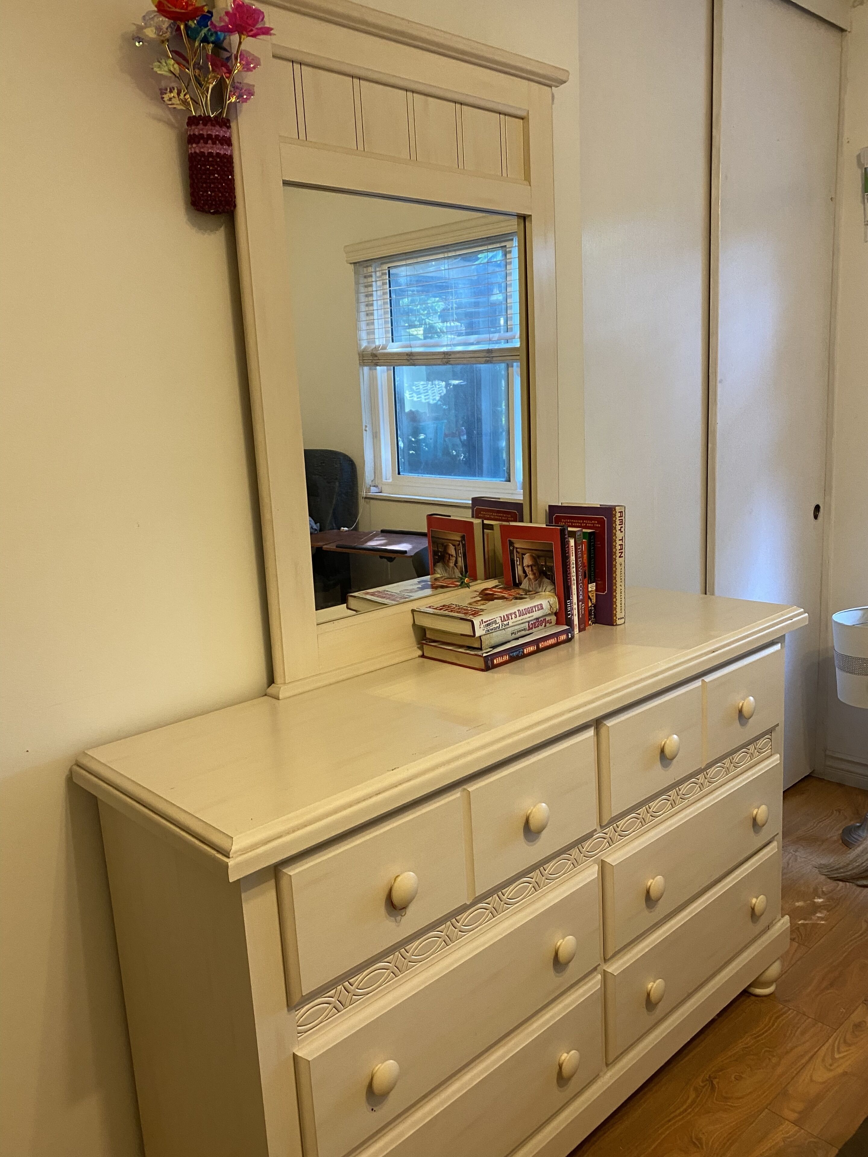 Une commode blanche avec un miroir au-dessus, une fenêtre avec des stores et un petit bureau avec une chaise.