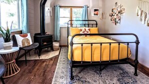 Un dormitorio acogedor con cama, una mesita, una silla, aire acondicionado instalado en la pared y una ventana con cortinas.