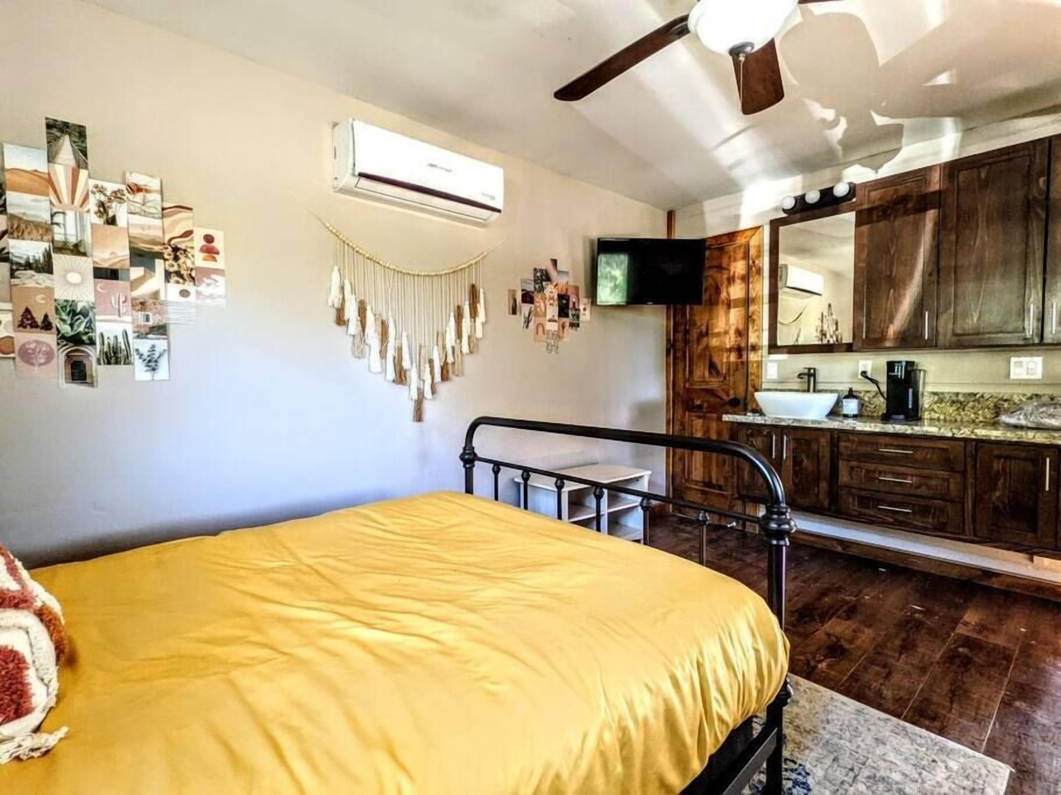 Une chambre à coucher comprenant un lit, un ventilateur de plafond, un climatiseur, un téléviseur fixé au mur et un coin cuisine avec des meubles en bois et un évier.
