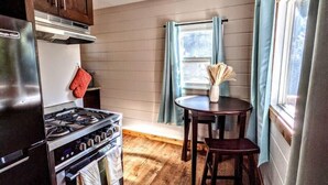 Cocina compacta con estufa, horno y armarios de madera. Hay una pequeña mesa de comedor con sillas y una ventana con cortinas.