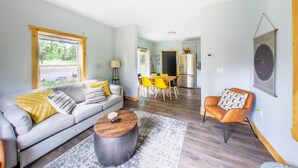 Ein modernes Wohnzimmer mit Sofa, Couchtisch und Sesseln. Es gibt einen Essbereich mit gelben Stühlen und eine Küche mit Kühlschrank.