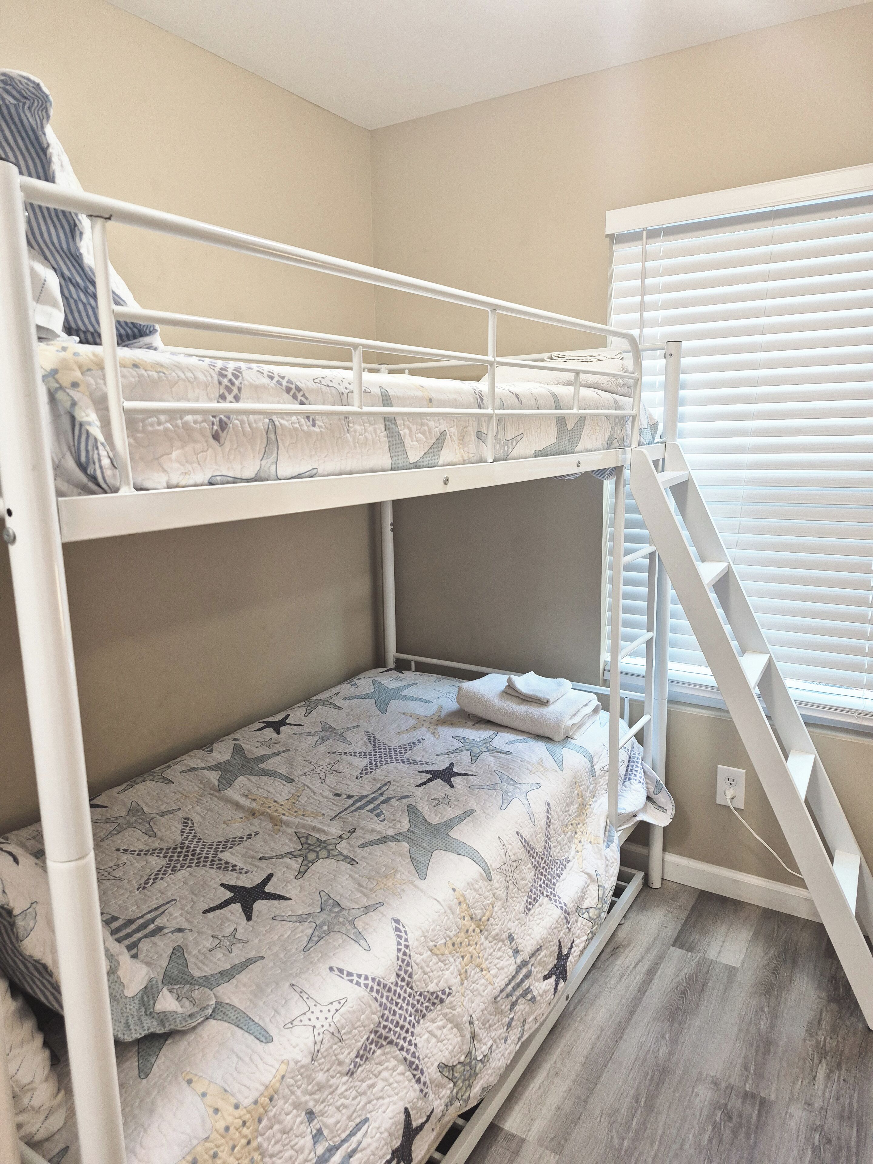 A bunk bed room with a star-patterned bedspread, a white metal ladder, and a window with blinds.