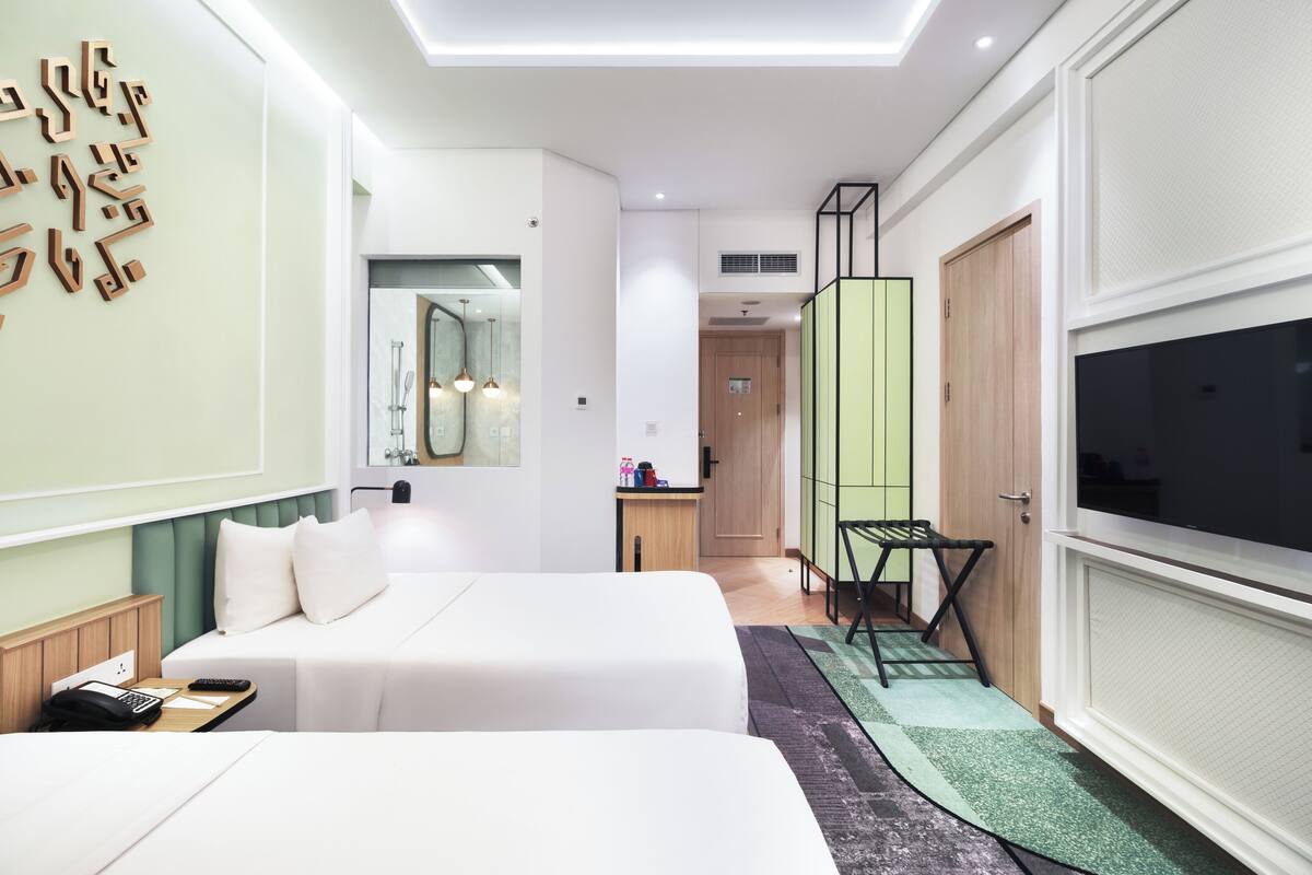 Quarto de hotel moderno com duas camas, televisão, escrivaninha com cadeira e banheiro visível através de uma porta aberta.