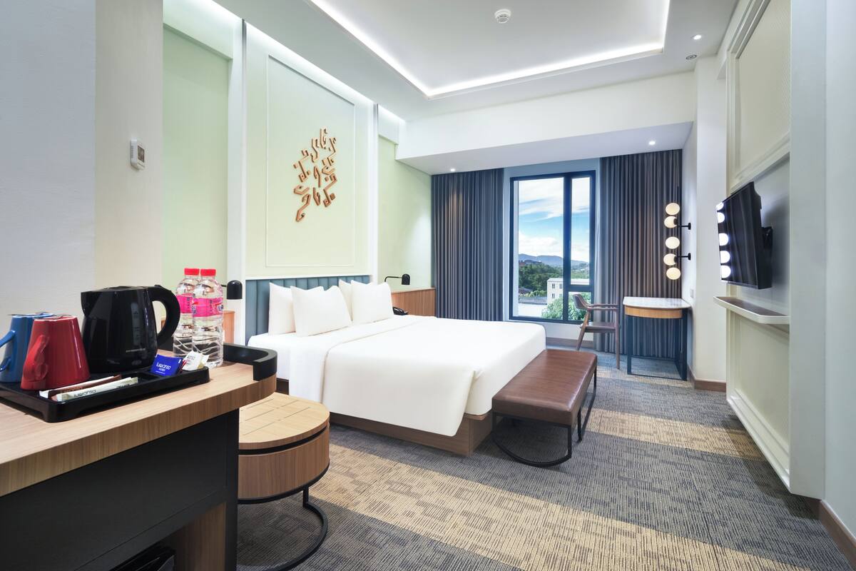 Quarto de hotel com uma cama grande, escrivaninha com cafeteira, uma cadeira, uma mesinha e televisão.