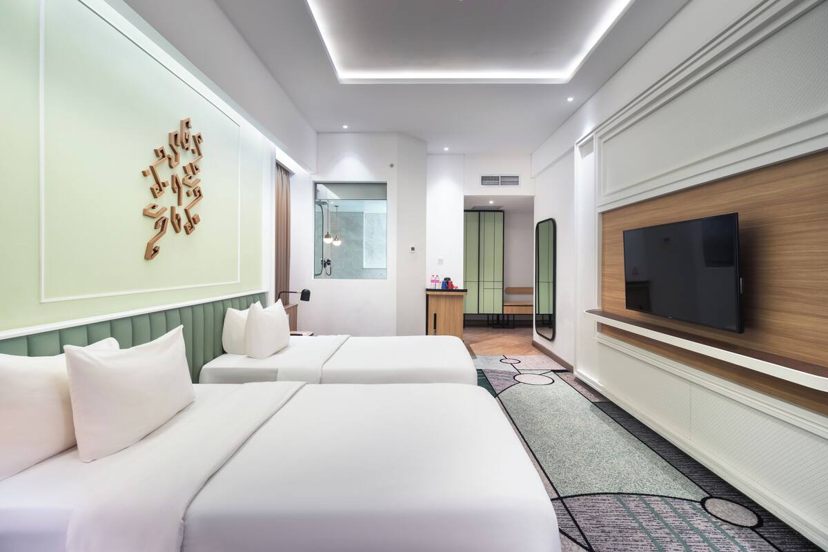 Quarto de hotel com duas camas, televisão, espelho e banheiro visível através de uma porta de vidro.
