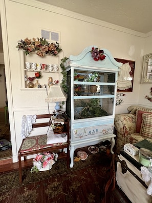 Una habitación de estilo vintage con un arreglo floral en un estante, una silla de madera con encaje y una mesita con un florero.