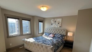 A bedroom with a tufted headboard bed, a nightstand with a lamp, and a window with a view of buildings. - Three Bedroom House,  sleeps 7 (Edmonton)