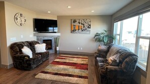 A living room with two patterned sofas, a fireplace, a flat-screen TV, a large window, and a cityscape painting. - Three Bedroom House,  sleeps 7 (Edmonton)