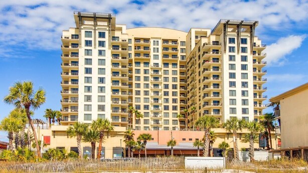 A multi-story hotel with balconies, located near a beach with palm trees.
