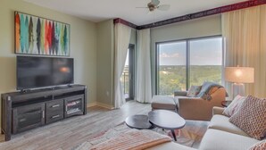 A modern living room with a flat-screen TV, a sofa, a coffee table, and a view of the outdoors through large windows.