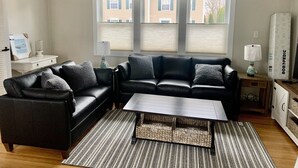 A living room with a black leather sofa, a coffee table, and a striped rug. There are two windows with blinds, a white side table, and a lamp. - Beach Ready 3-bdrm house, Short Walk to Private Beach and Park, Lots of Parking (Niantic)