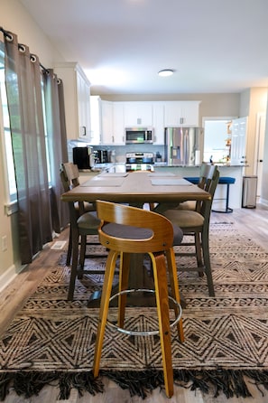 A dining area with a wooden table and chairs, a patterned rug, and a view into a kitchen with white cabinets and appliances.