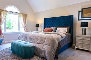 A bedroom with a tufted headboard, a large window with curtains, a bedside table with lamps, and a matching ottoman.