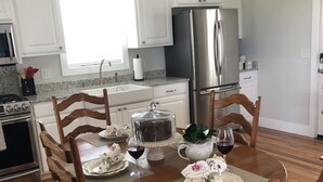A dining area with a wooden table set for four, surrounded by wooden chairs. A kitchen with white cabinets and stainless steel appliances is visible in the background. - The White Magnolia Cottage (Round Top)