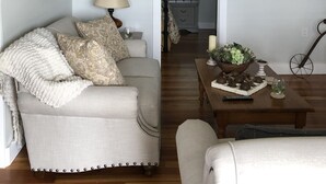 A living room with a sofa, a coffee table, and a lamp. There is a hallway leading to a kitchen. - The White Magnolia Cottage (Round Top)