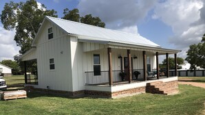 A white single-story building with a covered porch and a brick foundation. - The White Magnolia Cottage (Round Top)