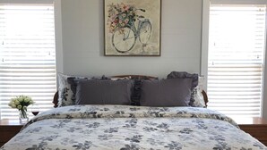 A bedroom with a floral bedspread, a framed artwork of a bicycle, and a vase of flowers on the nightstand. - The White Magnolia Cottage (Round Top)
