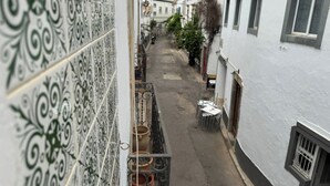 A narrow street with white buildings, a tiled wall, and a small balcony with potted plants.