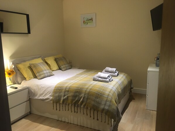 A neatly made bed with a plaid blanket and pillows, a bedside table with a lamp and a vase of sunflowers, and a small cabinet to the right. - Private Room - Sleeps 2 - Private Entry - Parking (Solihull)