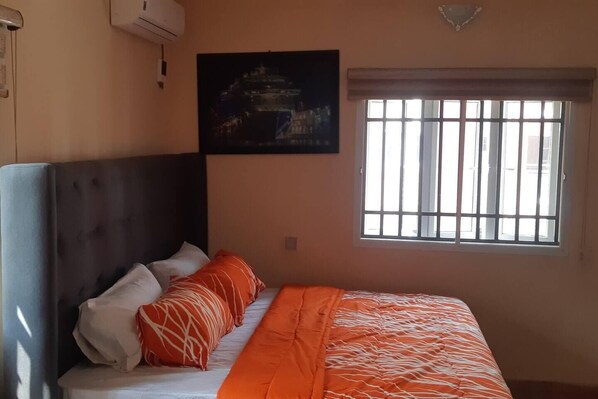 A bedroom with a bed, pillows, a window with bars, and a painting on the wall.