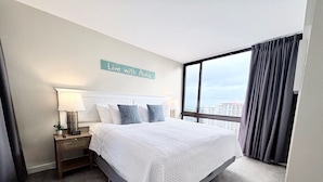 A hotel room with a large bed, a nightstand with a lamp, a view of the city through a large window, and a sign on the wall that says "Live with Aloha". - Renovated - Diamond Head and Ocean Views! - FREE Parking - 1 Block to Beach (Honolulu)