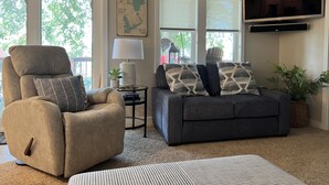A living room with a beige recliner, a dark gray sofa, a lamp, a map on the wall, and a TV mounted above the sofa. - Lakeview Cabin (Spirit Lake)