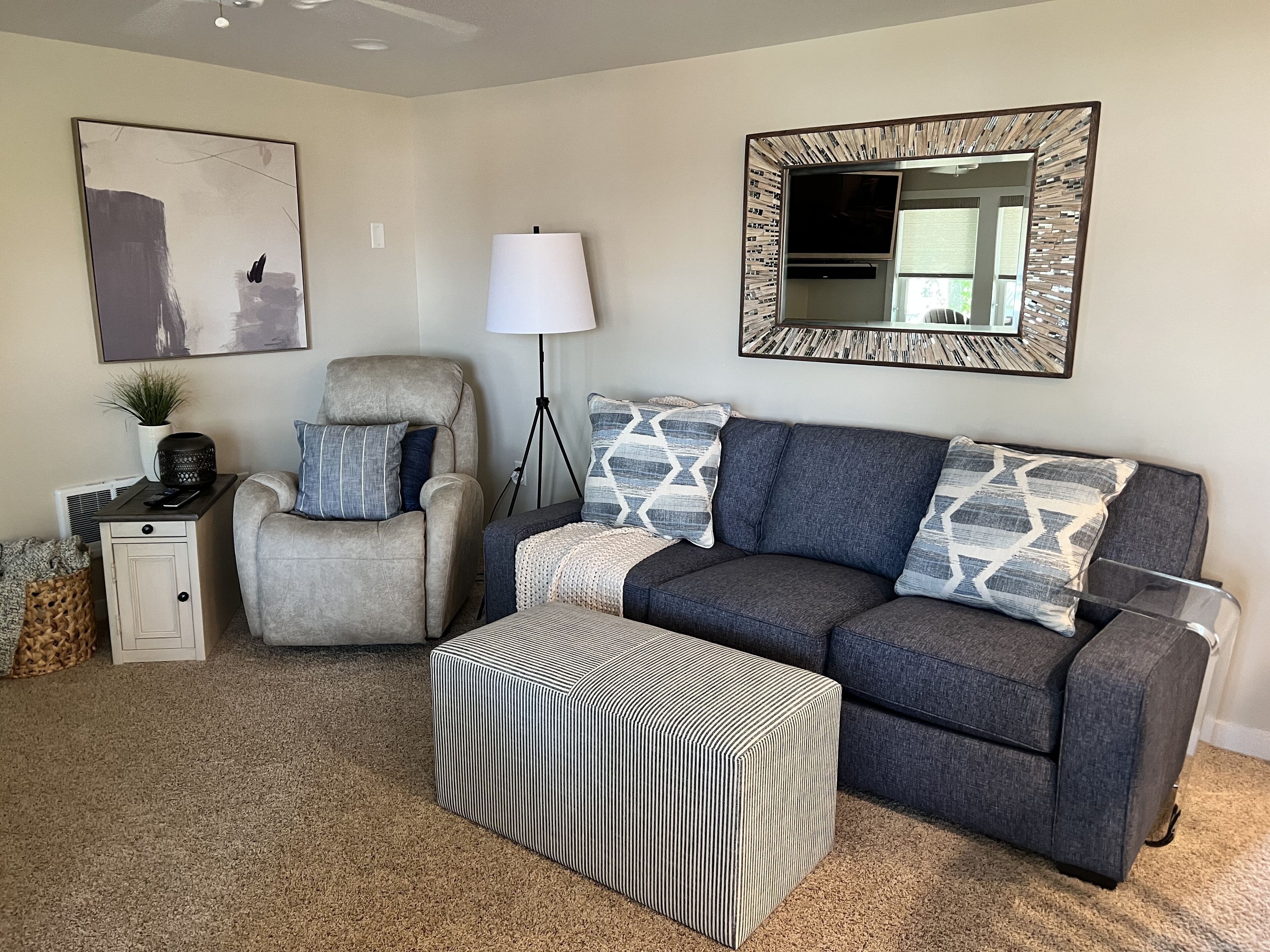 A living room with a grey sofa, a striped ottoman, a floor lamp, and a framed mirror.