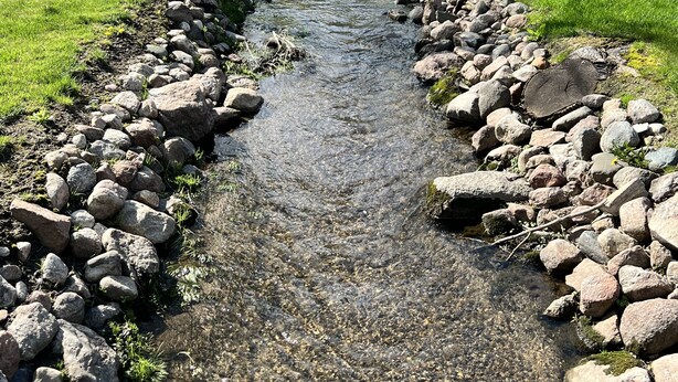 A clear stream with a stone border, surrounded by grass and trees, leading to a house with a dark roof and white trim. - Lakeview Cabin (Spirit Lake)