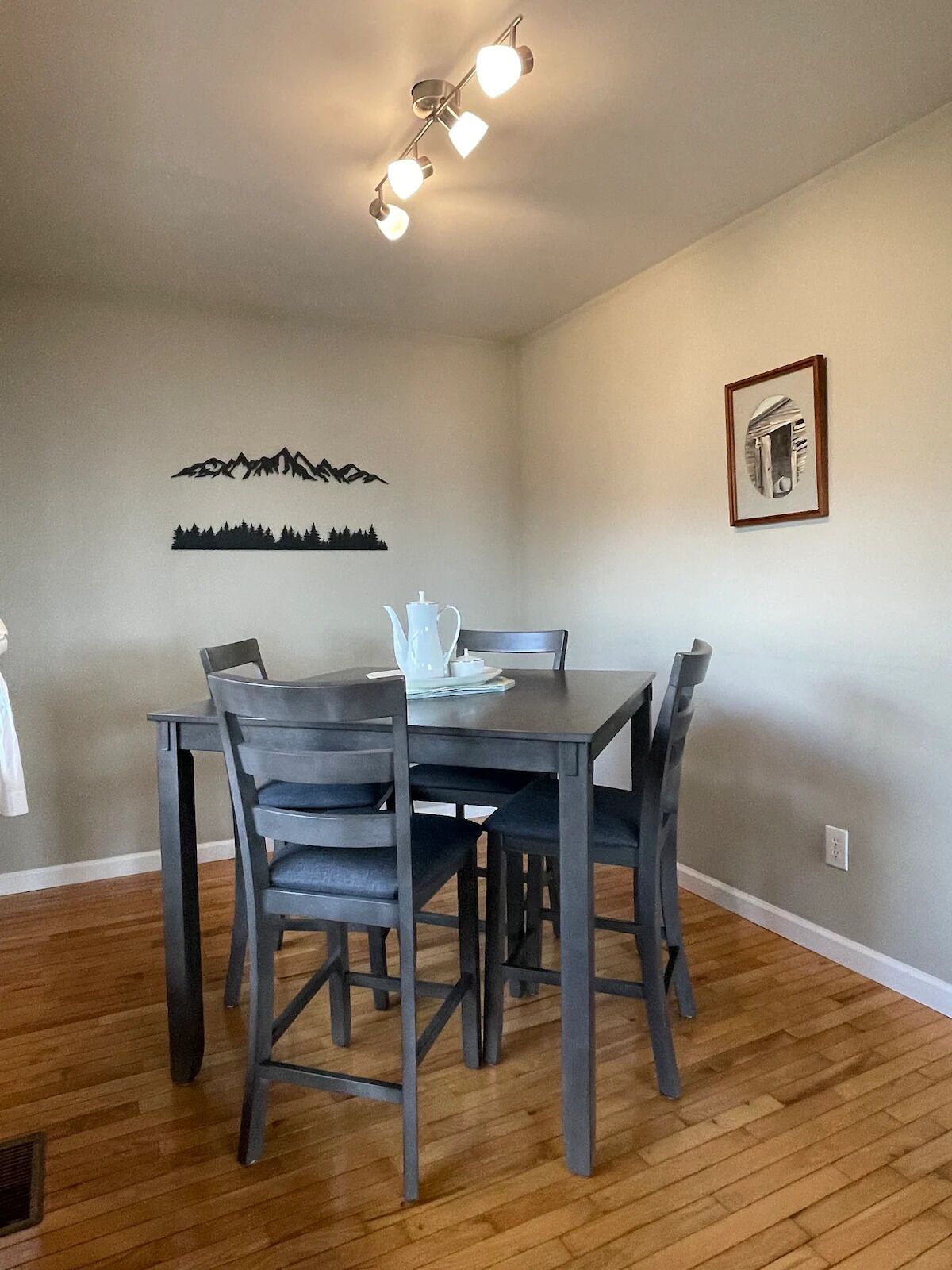 A dining area with a wooden table and chairs, a ceiling light fixture, and wall art depicting mountains and trees.