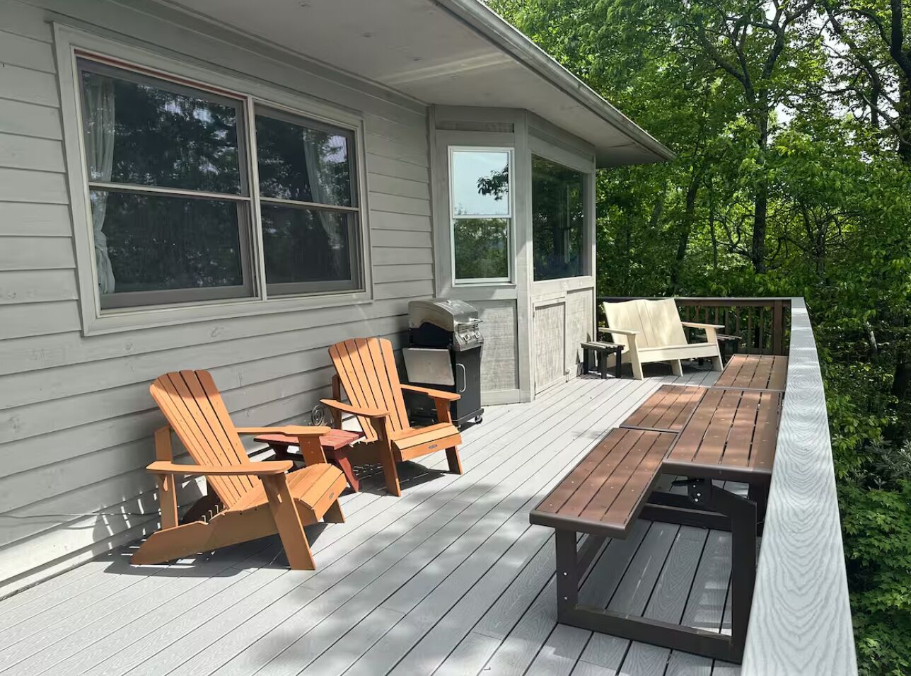 En træterrasse med Adirondack-stole og et picnicbord, omgivet af et hus med store vinduer og en overdækket terrasse.