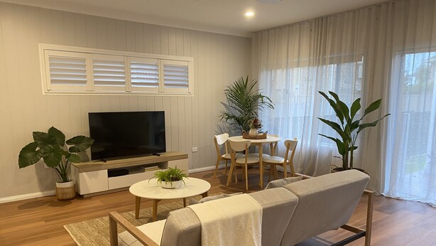 A modern living room with a flat-screen TV, a sofa, a dining table with chairs, and potted plants.