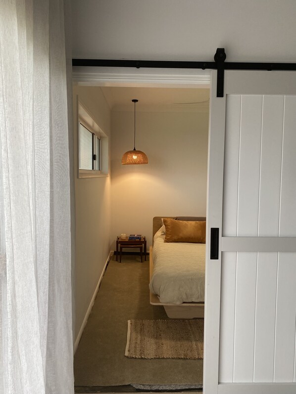 A bedroom with a bed, a bedside table with books and a cup, a hanging light fixture, and a door with a black frame.