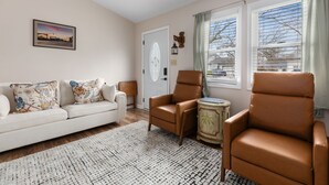 Ein Wohnzimmer mit einer Couch, zwei Sesseln, einem gemusterten Teppich, einer Holzkommode und einer Tür mit geometrischem Muster.