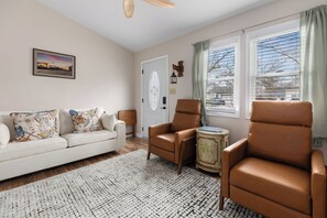 Un salon comprenant un canapé, deux fauteuils, une petite table d’appoint, un tapis à motifs, une porte ornée d’un panneau décoratif et une fenêtre avec des stores.