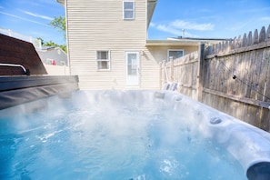 A hot tub in a backyard with a wooden fence, a house, and a clear sky.