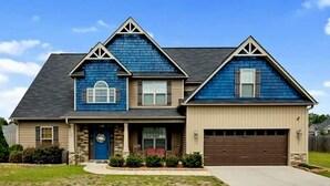 A two-story house with a dark blue exterior, a brown garage door, and a front porch with a red bench. - 🌟"Home-Away-From-Home" Beauty; Kid, Baby & Dog-Friendly; Veteran-Owned🌟 (Sanford)