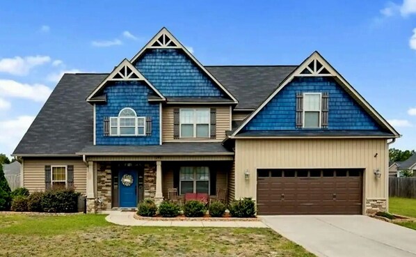 A two-story house with a dark blue exterior, a brown garage door, and a front porch with a red bench. - 🌟"Home-Away-From-Home" Beauty; Kid, Baby & Dog-Friendly; Veteran-Owned🌟 (Sanford)