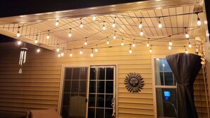 An outdoor patio area with string lights, a sliding glass door, a sun-shaped wall decoration, and a window with curtains. - 🌟"Home-Away-From-Home" Beauty; Kid, Baby & Dog-Friendly; Veteran-Owned🌟 (Sanford)