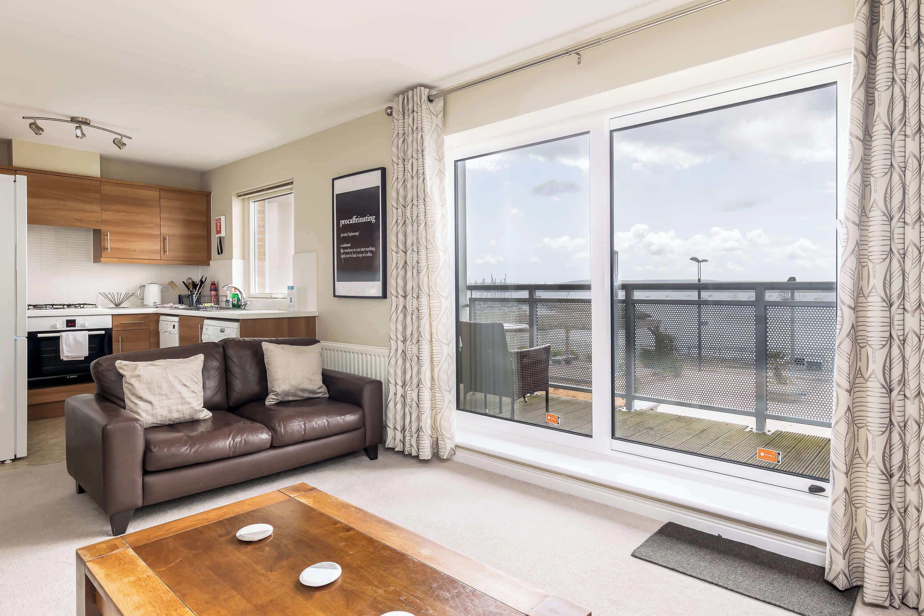 Un salon moderne avec un canapé en cuir marron, une table basse en bois et une grande porte-fenêtre coulissante donnant sur un balcon avec vue sur la mer.
