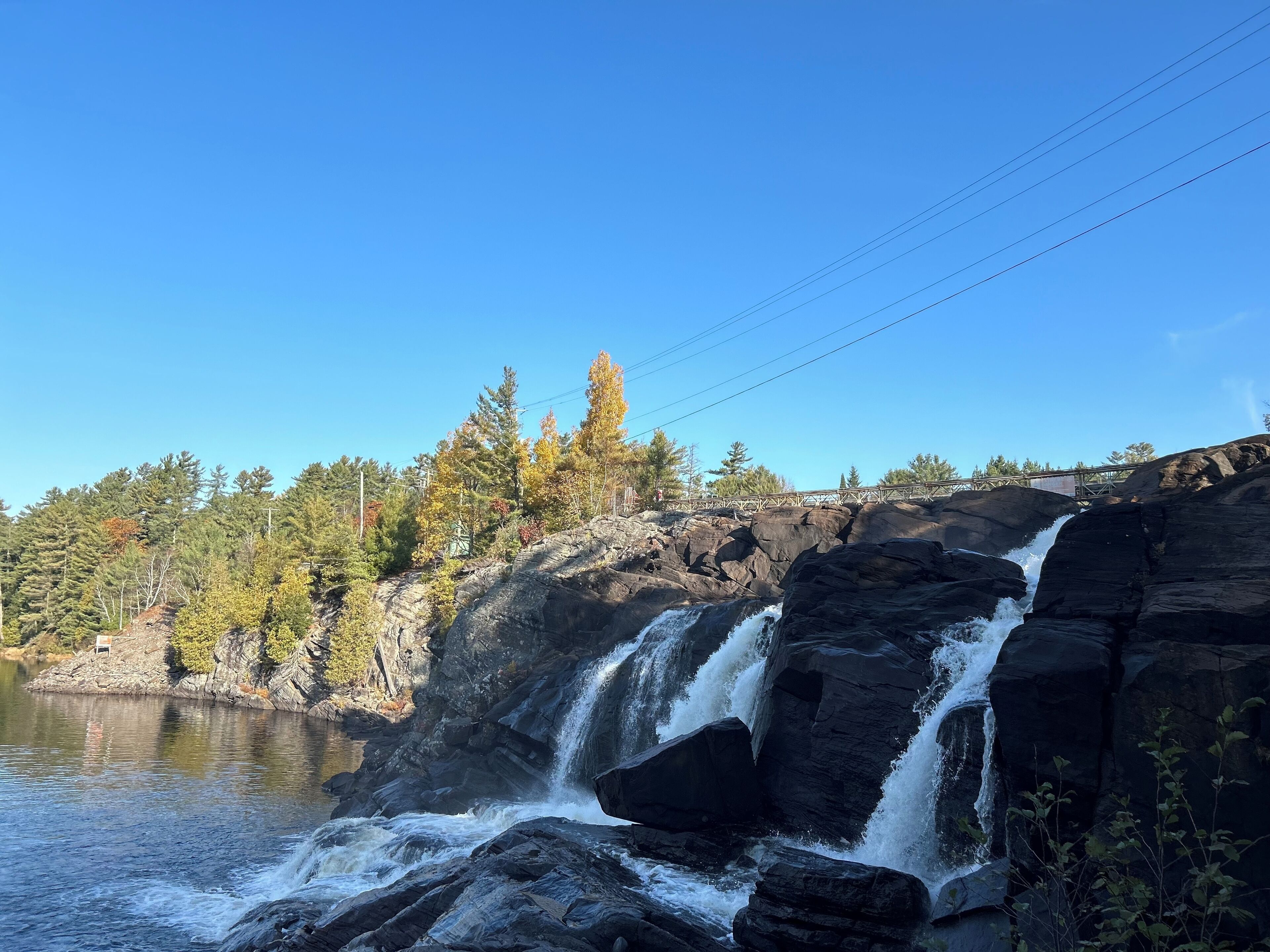 A waterfall cascading over dark rocks into a calm river, surrounded by trees and a clear blue sky.