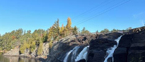 A waterfall cascading over dark rocks into a calm river, surrounded by trees and a clear blue sky.