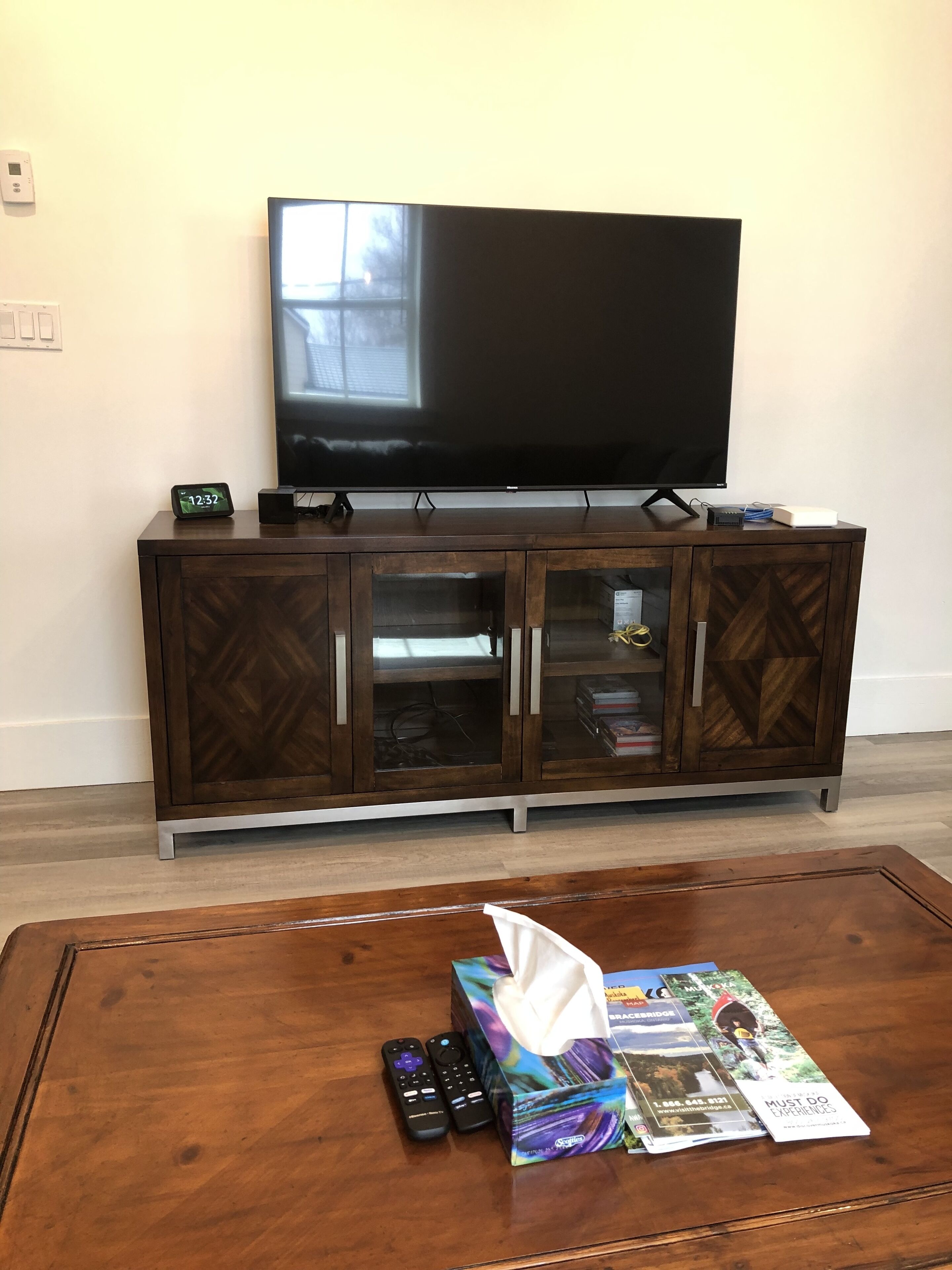 A living room with a TV on a wooden cabinet, a coffee table with remote controls and magazines, and a tissue box.