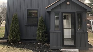 Une maison à deux étages, à l’extérieur de couleur gris foncé, avec un porche à l’avant doté d’une petite fenêtre, et deux arbres à feuilles persistantes dans le jardin.