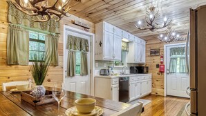 A wooden interior with a dining table set for two, a kitchen with white cabinets, and a chandelier. - Cozy Cabin Hot tub Firepit Game Room Pool Table Gas Fireplace 5 min to GB-PF (Sevierville)