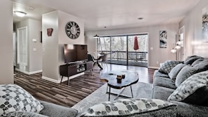 Een moderne woonkamer met een bank, salontafel en televisie. Er is een eethoek met stoelen en een tafel, en een balkon met een balustrade.