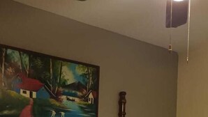 A neatly made bed with a wooden headboard, a painting of a landscape on the wall, and a ceiling fan. - Suit Rivas 132 Gran Pacifica Resort (Villa El Carmen)