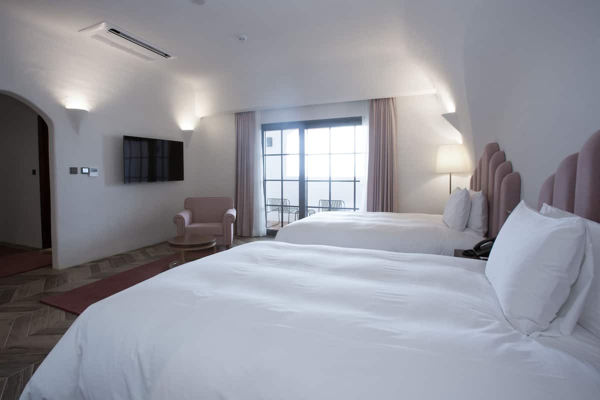 A hotel room with two beds, a TV mounted on the wall, a chair, and a balcony with a view.