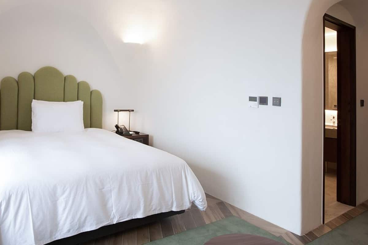 A neatly made bed with a green headboard, a bedside table with a lamp, and a door leading to another room.
