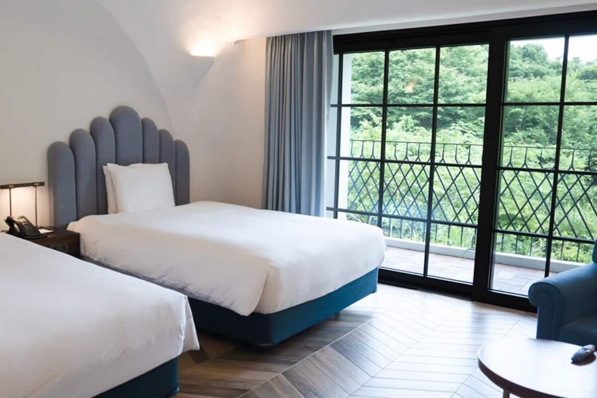A hotel room with two beds, a desk, a chair, and a large window with a view of greenery.
