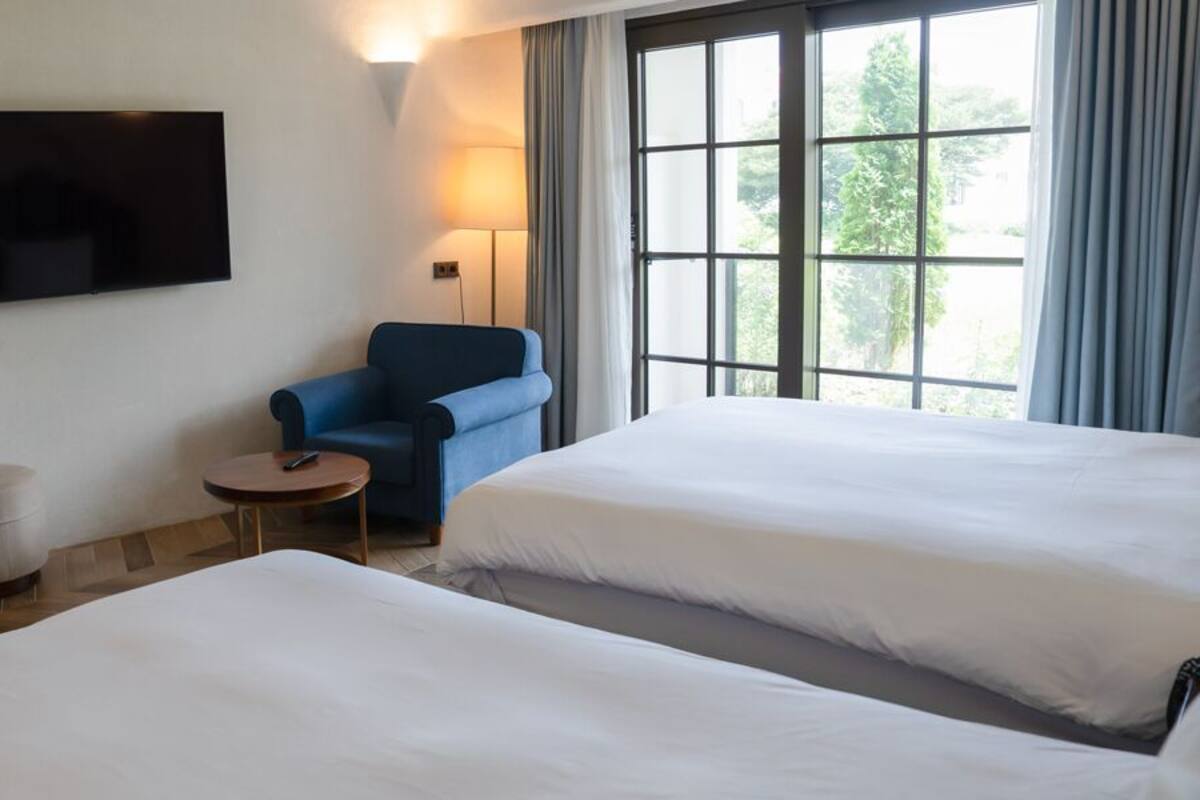 A hotel room with two beds, a desk with a chair, a TV, and a window with curtains.
